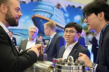 Several participants standing at an exhibition booth, examining a large metallic technical component together. A large ship illustration is visible in the background.