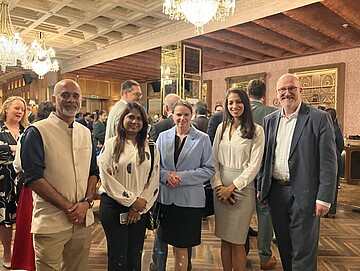Eine Gruppe von fünf Personen steht zusammen in einem elegant eingerichteten Veranstaltungsraum mit Kronleuchtern, Holzdecke und dekorativen Wänden. Hinter ihnen sind zahlreiche weitere Gäste zu sehen, die sich unterhalten. Die Personen in der Vorderreihe stehen locker nebeneinander und tragen formelle oder halbformelle Kleidung, passend zu einem Empfang oder Networking‑Event.