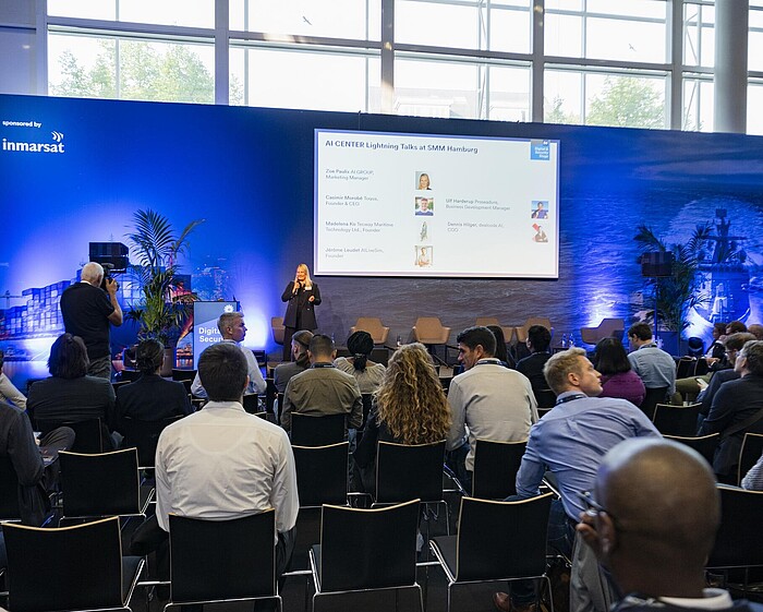 Blick von hinten auf das Publikum, während eine Speakerin vorne auf der Bühne steht. Auf der Leinwand ist die Aufschrift ‚AI CENTER Lightning Talks at SMM Hamburg‘ zu sehen.
