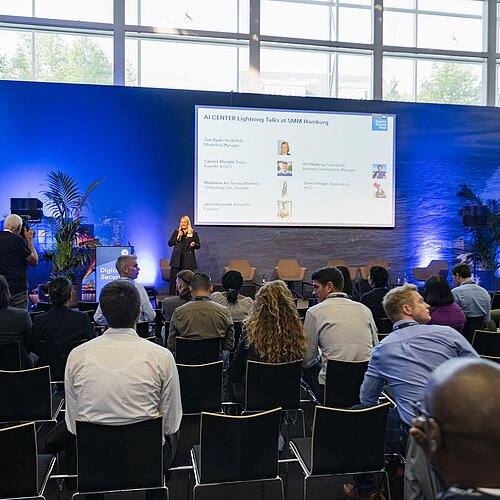 Blick von hinten auf das Publikum, während eine Speakerin vorne auf der Bühne steht. Auf der Leinwand ist die Aufschrift ‚AI CENTER Lightning Talks at SMM Hamburg‘ zu sehen.