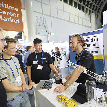 Besucherinnen und Besucher informieren sich an einem Unternehmensstand auf der SMM auf dem Maritime Career Market; auf dem Tresen sind ein Laptop und ein Modellkran zu sehen.
