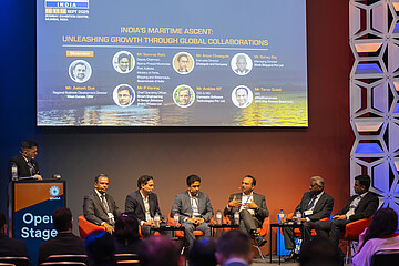 A panel discussion taking place on a conference stage, with several speakers seated in front of an audience. A large screen in the background displays the topic of maritime growth and international collaboration.