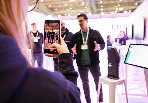 Blurry picture with focus on a womans phone, taking a video of two people talking.