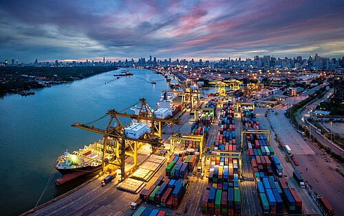 Aerial view of port with crane loading containers onto ship, against backdrop of Bangkok skyline at night.