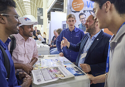 Ausstellende unterhalten sich mit Besuchenden an einem Pult.