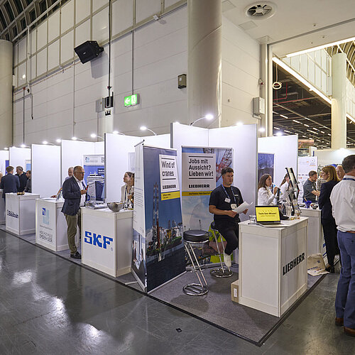 In einer Ausstellungshalle sind mehrere Stände in einer Reihe angeordnet. Ausstellende sind an den Ständen zu sehen, während Besuchende den danebenliegenden Gang entlanggehen.