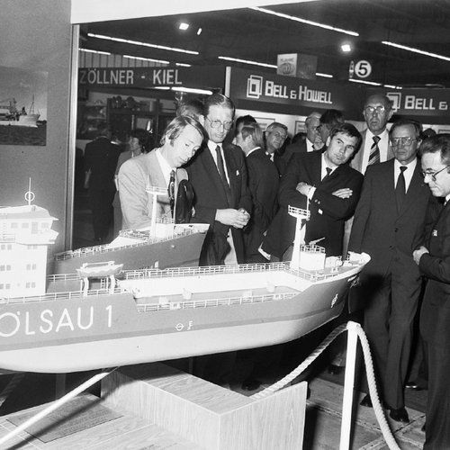 Multiple fair visitors, who are looking at the miniature ship "Öslau 1". There's more exhibition stands in the background.