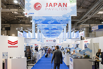 View through the Japan Pavilion in an exhibition hall, showing a central blue carpet leading past maritime industry booths. Overhead banners with wave patterns and the text “Japan Pavilion” hang from the ceiling.