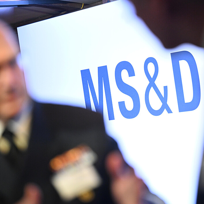 Unscharfes Foto einer Veranstaltungsszene: Im Vordergrund steht ein deutlich sichtbares blaues „MS&D“-Logo auf einer hellen Fläche, während im Hintergrund eine Person in dunkler Kleidung mit Abzeichen stark verschwommen zu sehen ist.