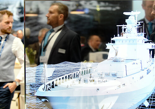 Ein weißes Schiff ist auf einer Glasscheibe abgebildet. Menschen laufen im Hintergrund bei der Messe umher. 