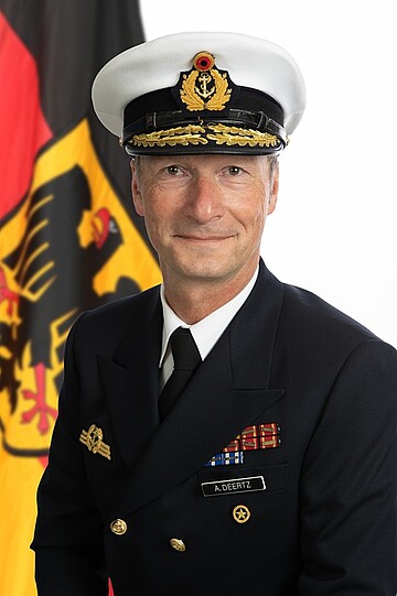 „Person in dunkler Uniform und weißer Kopfbedeckung vor einer Flagge; auf der Uniform ist ein Namensschild mit der Aufschrift ‚A. DEERTZ‘ zu sehen.“