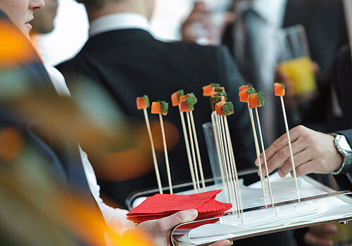Close-up of a tray with long sticks, holding a small treat at the end. A crowd is visible all around.