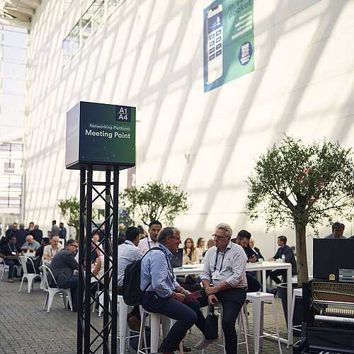 Ein Meeting Point auf der Hamburg Messe, im Hintergrund sind Tische und Stühle zu sehen, an denen Besuchende sitzen und sich unterhalten.