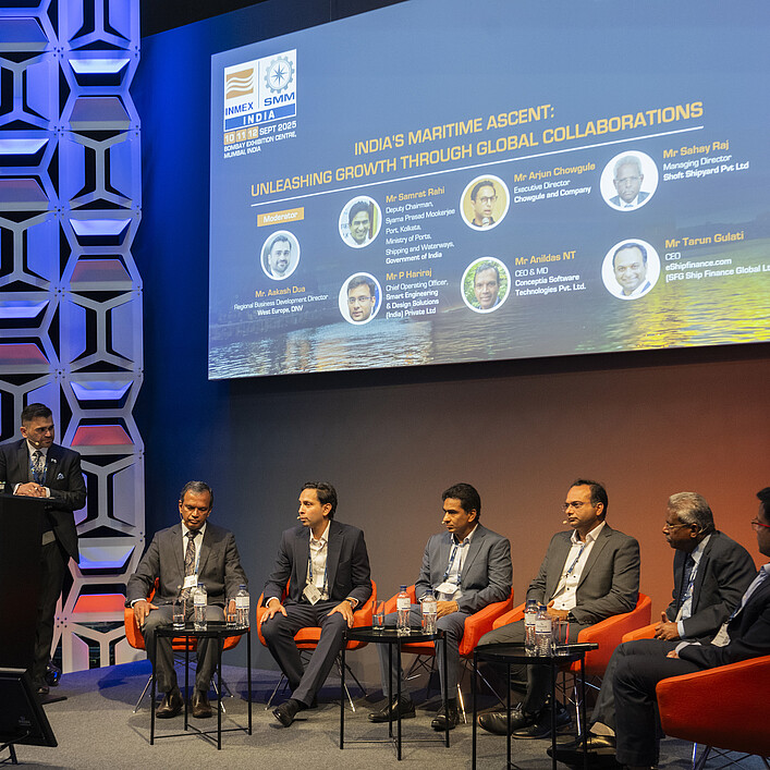 Panel discussion on a conference stage. One person stands at a lectern labeled “Open Stage” while six other participants are seated in orange chairs. A large screen in the background displays information about a maritime industry event in India. The stage is lit in blue and orange tones and framed by decorative wall panels.