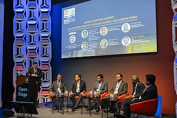 Eine Podiumsdiskussion auf einer Bühne mit sieben Personen, von denen eine am Rednerpult steht und sechs in einer Reihe auf orangefarbenen Sesseln sitzen. Hinter ihnen zeigt eine große Leinwand den Titel der Veranstaltung zum Thema maritime Entwicklung und internationale Zusammenarbeit. Die Bühne ist modern ausgeleuchtet und hat eine geometrische Wandgestaltung. Das Publikum ist nicht sichtbar.