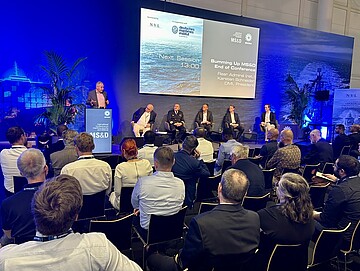 Konferenzraum mit Publikum; auf einer Bühne sitzen mehrere Personen in einer Podiumsrunde, während eine Person am Rednerpult spricht. Im Hintergrund ist eine große Leinwand mit Präsentationsfolien zu sehen.