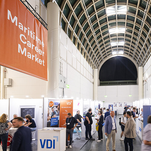 Eine Ausstellungshalle mit sichtbaren Ständen, Ausstellenden und Besuchenden. Im Vordergrund und Hintergrund ist der Hauptgang der Halle erkennbar, der die Stände miteinander verbindet.