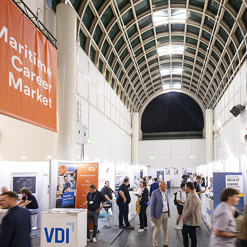 Eine Ausstellungshalle mit sichtbaren Ständen, Ausstellenden und Besuchenden. Im Vordergrund und Hintergrund ist der Hauptgang der Halle erkennbar, der die Stände miteinander verbindet.