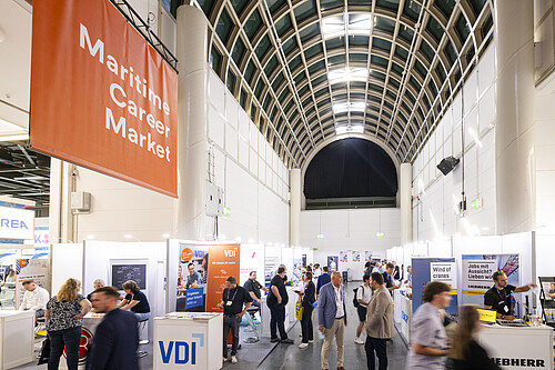 Eine Ausstellungshalle mit sichtbaren Ständen, Ausstellenden und Besuchenden. Im Vordergrund und Hintergrund ist der Hauptgang der Halle erkennbar, der die Stände miteinander verbindet.