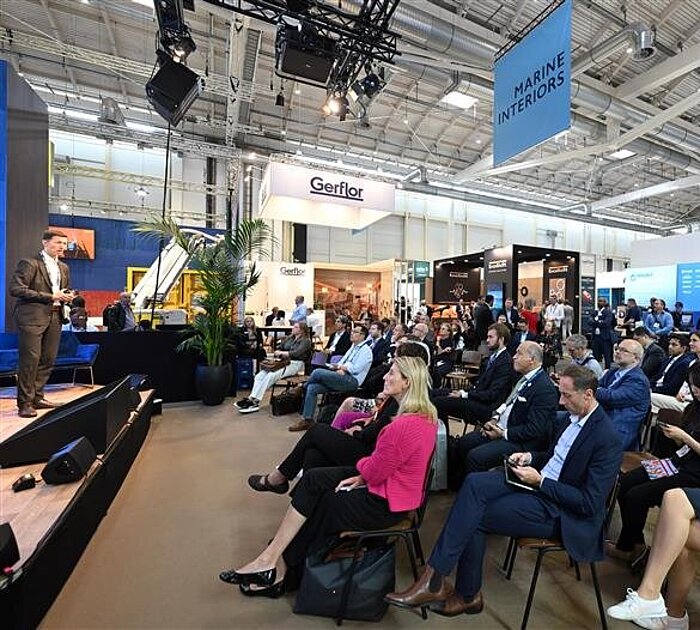 Spectators look at a stage and listen to a lecture. A sign reading "Marine Interiors" hangs above them.
