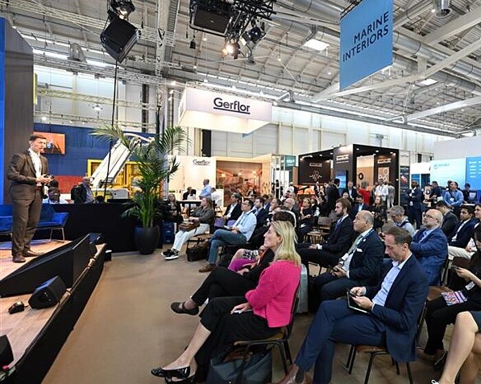 Spectators look at a stage and listen to a lecture. A sign reading "Marine Interiors" hangs above them.