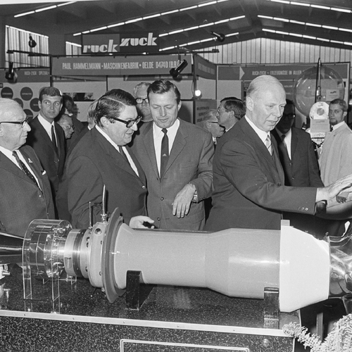  Historical image of a group of trade fair visitors looking at a machine part on display. Other trade fair stands and visitors can be seen in the background.